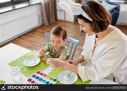 holidays, family and people concept - happy mother and little baby daughter coloring easter eggs with watercolors at home. mother with child coloring easter eggs at home
