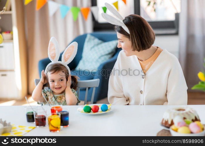 holidays, family and people concept - happy mother and little baby daughter in bunny ears dyeing easter eggs at home. mother with child dyeing easter eggs at home