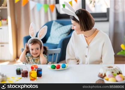 holidays, family and people concept - happy mother and little baby daughter in bunny ears dyeing easter eggs at home. mother with child dyeing easter eggs at home