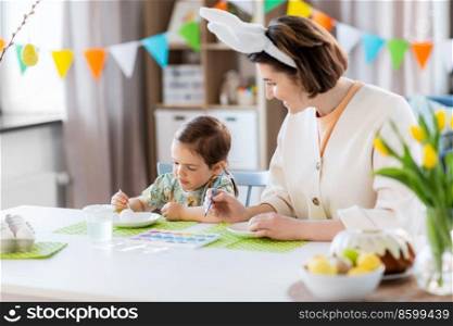 holidays, family and people concept - happy mother and little baby daughter coloring easter eggs with watercolors at home. mother with child coloring easter eggs at home