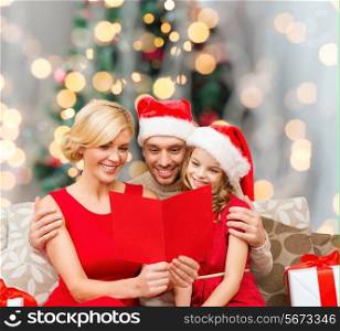 holidays, family and people concept - happy mother, father and little girl in santa helper hats with gift boxes reading geeting card over living room and christmas tree background