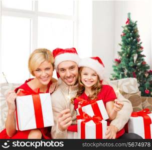 holidays, family and people concept - happy mother, father and little girl in santa helper hats with gift boxes and sparklers over living room and christmas tree background