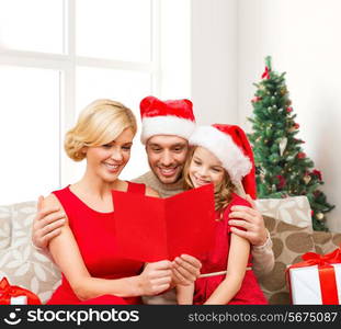 holidays, family and people concept - happy mother, father and little girl in santa helper hats with gift boxes reading geeting card over living room and christmas tree background