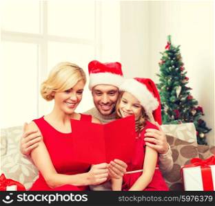 holidays, family and people concept - happy mother, father and little girl in santa helper hats with gift boxes reading geeting card over living room and christmas tree background