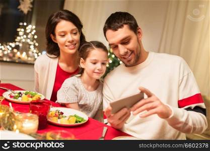 holidays, family and technology concept - happy mother, father and little daughter with smartphone having christmas dinner at home. family with smartphone having christmas dinner