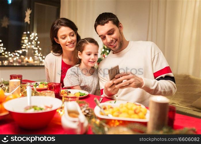 holidays, family and technology concept - happy mother, father and little daughter with smartphone having christmas dinner at home. family with smartphone having christmas dinner