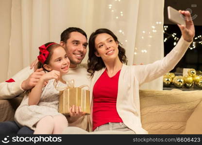 holidays, family and technology concept - happy mother, father and little daughter with christmas gift taking selfie by smartphone at home. happy family with christmas present at home