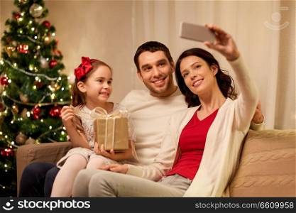holidays, family and technology concept - happy mother, father and little daughter with christmas gift taking selfie by smartphone at home. happy family with christmas present at home