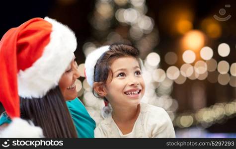 holidays, family, childhood and people concept - happy mother and little daughter in santa hats over christmas lights background. mother and daughter in santa hats at christmas