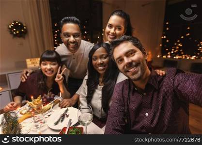holidays, friendship and celebration concept - multiethnic group of happy friends having christmas dinner at home and taking selfie. friends taking selfie on christmas dinner at home