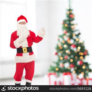 holidays, gesture and people concept - man in costume of santa claus pointing fingers over living room and christmas tree background