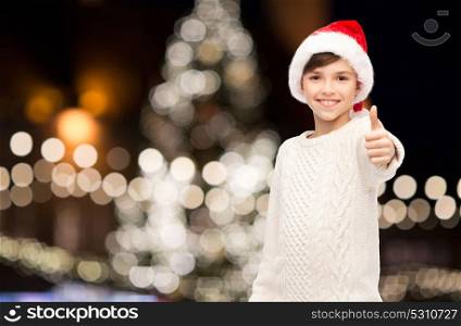 holidays, gesture, childhood and people concept - smiling happy boy in santa hat showing thumbs up over christmas tree lights background. boy in santa hat showing thumbs up at christmas