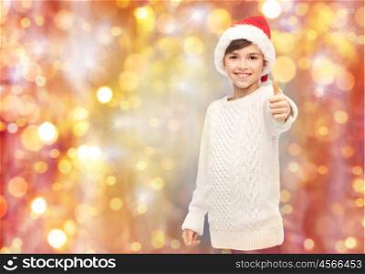 holidays, gesture, christmas, childhood and people concept - smiling happy boy in santa hat showing thumbs up over lights background