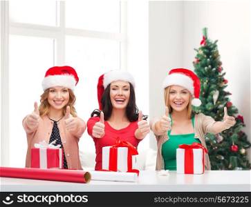 holidays, gesture, decoration and people concept - smiling women in santa helper hats with decorating paper and gift boxes showing thumbs up over living room and christmas tree background