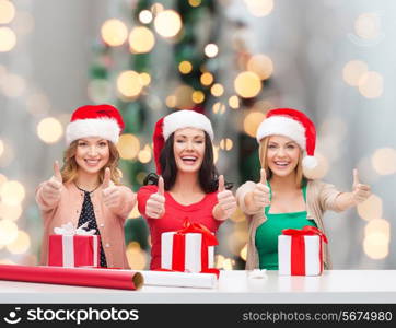 holidays, gesture, decoration and people concept - smiling women in santa helper hats with decorating paper and gift boxes showing thumbs up over christmas tree background