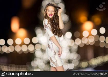 holidays, hairstyle and people concept - happy young woman or teen girl in fancy dress with sequins touching long wavy hair over christmas tree lights background. happy young woman over christmas tree lights