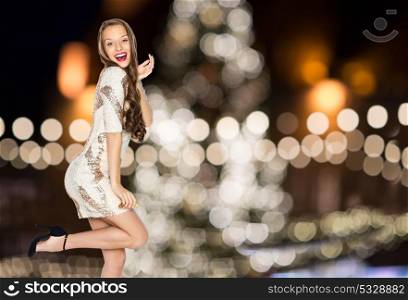 holidays, hairstyle and people concept - happy young woman or teen girl in fancy dress with sequins posing over christmas tree lights background. happy woman posing over christmas tree lights