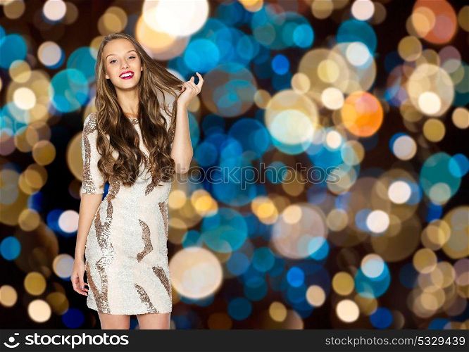 holidays, hairstyle and people concept - happy young woman or teen girl in fancy dress with sequins touching long wavy hair over festive lights background. happy young woman over festive lights