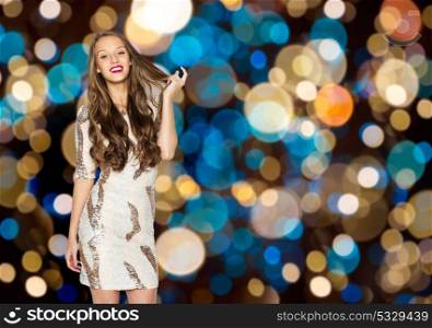 holidays, hairstyle and people concept - happy young woman or teen girl in fancy dress with sequins touching long wavy hair over festive lights background. happy young woman over festive lights