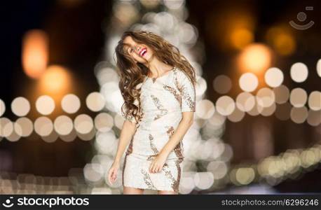 holidays, hairstyle and people concept - happy young woman or teen girl in fancy dress dancing over christmas tree lights background. happy young woman over christmas tree lights