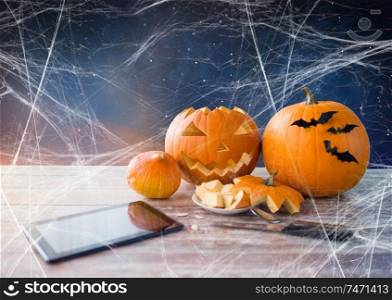 holidays, halloween and technology concept - jack-o-lantern or carved pumpkin, tablet pc computer, knife on wooden table and spiderweb over starry night sky background. halloween jack-o-lantern, pumpkins and tablet pc