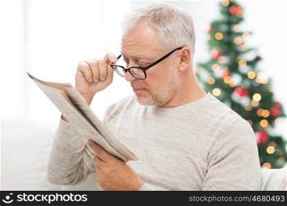 holidays, information, people, vision and mass media concept - senior man in glasses reading newspaper at home over christmas tree background