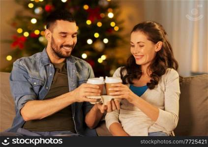 holidays, leisure and people concept - happy couple drinking takeaway tea or coffee at home in evening over christmas tree lights on background. happy couple drinking coffee at home on christmas