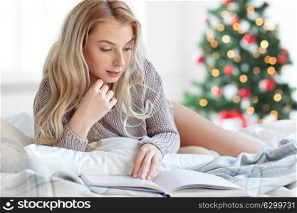 holidays, leisure and people concept - happy young woman reading book in bed at home bedroom over christmas tree background. young woman reading book in bed at christmas