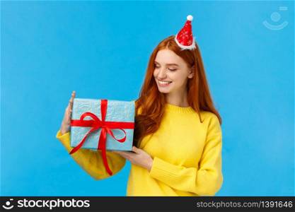 Holidays, love and family concept. Carefree tender and pretty redhead female holding cute wrapped box with gift, looking at present delighted, smiling happy, standing blue background joyful.. Holidays, love and family concept. Carefree tender and pretty redhead female holding cute wrapped box with gift, looking at present delighted, smiling happy, standing blue background joyful