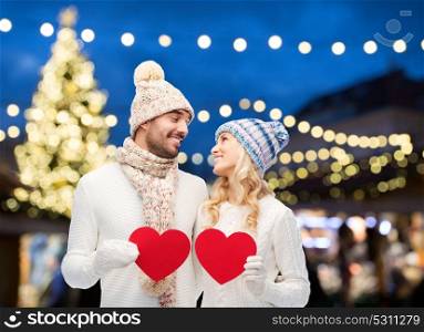 holidays, love and people concept - happy couple in winter hats holding red paper heart shapes over christmas tree lights background. couple with red hearts over christmas tree lights