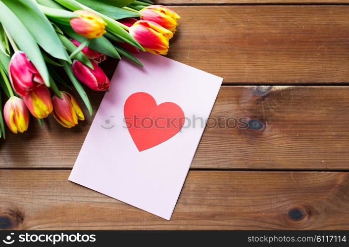 holidays, love and valentines day concept - close up of tulip flowers and greeting card with heart on wooden table