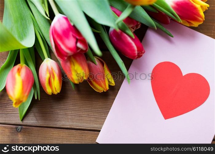 holidays, love and valentines day concept - close up of tulip flowers and greeting card with heart on wooden table