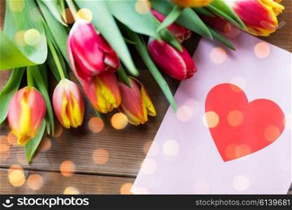 holidays, love and valentines day concept - close up of tulip flowers and greeting card with heart on wooden table over lights
