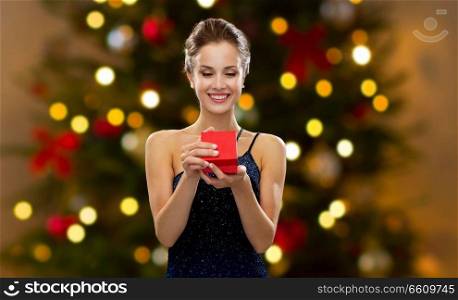 holidays, luxury and people concept - smiling woman in dress holding red gift box over christmas tree lights background. woman with red gift box over christmas tree lights