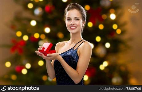 holidays, luxury and people concept - smiling woman in dress holding red gift box over christmas tree lights background. woman with red gift box over christmas tree lights