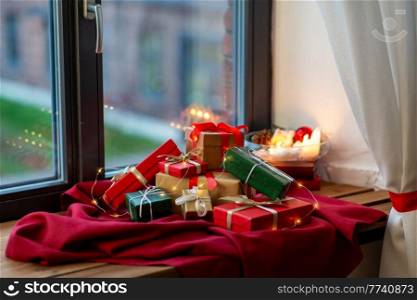 holidays, new year and celebration concept - heap of christmas gifts with electric garland on red tablecloth on window sill. christmas gifts on red tablecloth on window sill