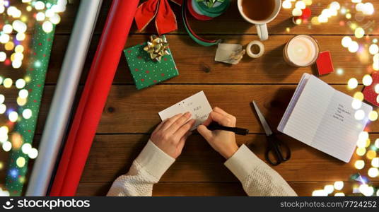 holidays, new year and christmas concept - hands packing gift box, attaching tag and writing receiver name on wooden table over festive lights. hands attaching name tag to christmas gift