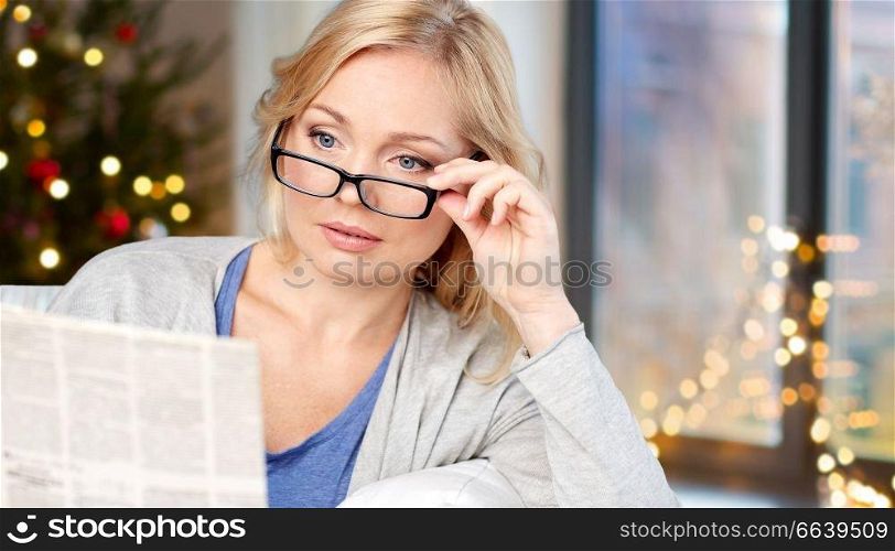 holidays, news and vision concept - middle aged woman in eyeglasses reading newspaper over christmas tree background at home. woman in glasses reading newspaper over christmas