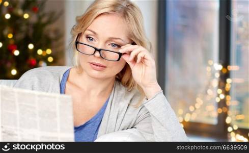 holidays, news and vision concept - middle aged woman in eyeglasses reading newspaper over christmas tree background at home. woman in glasses reading newspaper over christmas