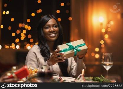 holidays, party and celebration concept - happy african american woman with gift box on christmas dinner at home. happy african woman with christmas gift at home