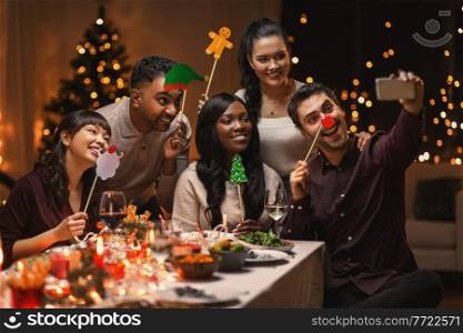 holidays, party and celebration concept - multiethnic group of happy friends having christmas dinner at home and taking selfie with smartphone. friends taking selfie on christmas dinner at home