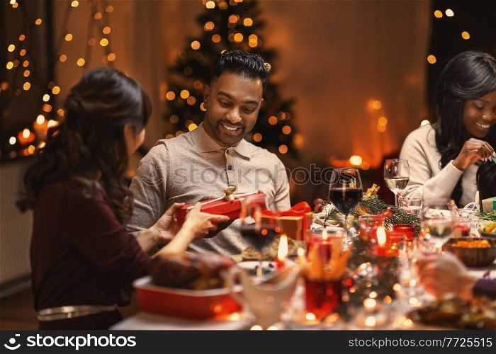 holidays, party and celebration concept - multiethnic group of happy friends having christmas dinner and giving presents at home. happy friends giving christmas presents at home