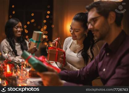 holidays, party and celebration concept - multiethnic group of happy friends having christmas dinner and opening presents at home. happy friends opening christmas presents at home