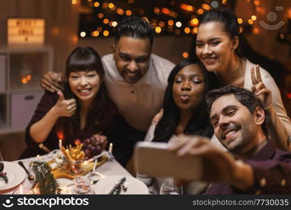 holidays, party and celebration concept - multiethnic group of happy friends having christmas dinner at home and taking selfie with smartphone. friends taking selfie on christmas dinner at home