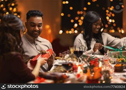 holidays, party and celebration concept - multiethnic group of happy friends having christmas dinner and giving presents at home. happy friends giving christmas presents at home