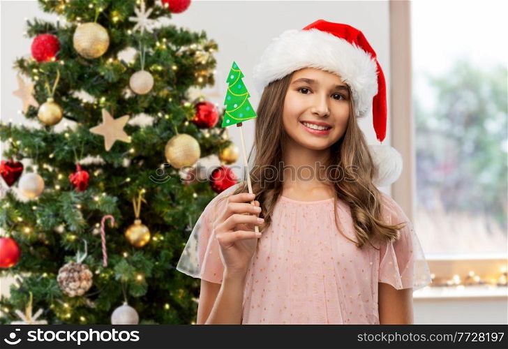holidays, party props and people concept - happy smiling teenage girl in santa helper hat accessory over christmas tree on background. happy teenage girl in santa hat on christmas