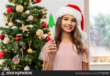 holidays, party props and people concept - happy smiling teenage girl in santa helper hat accessory over christmas tree on background. happy teenage girl in santa hat on christmas