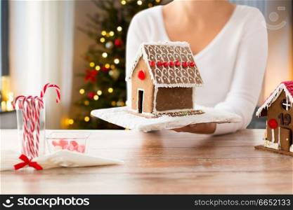 holidays, pastry and bakery concept - close up of woman holding and showing gingerbread house at home over christmas tree lights background. close up of woman with christmas gingerbread house