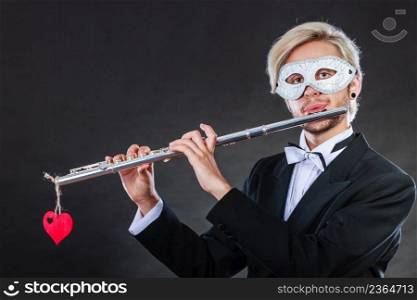 Holidays, people and celebration concept. Elegant young guy wearing suit white shirt bow tie and carnival venetian mask playing flute instrument on dark. man in carnival mask playing flute