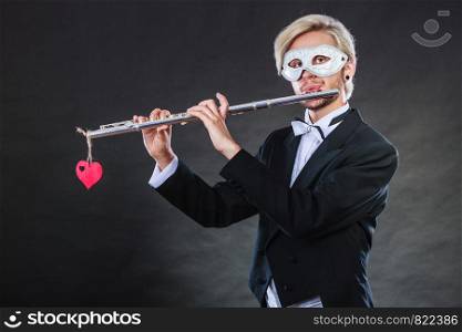 Holidays, people and celebration concept. Elegant young guy wearing suit white shirt bow tie and carnival venetian mask playing flute instrument on dark. man in carnival mask playing flute